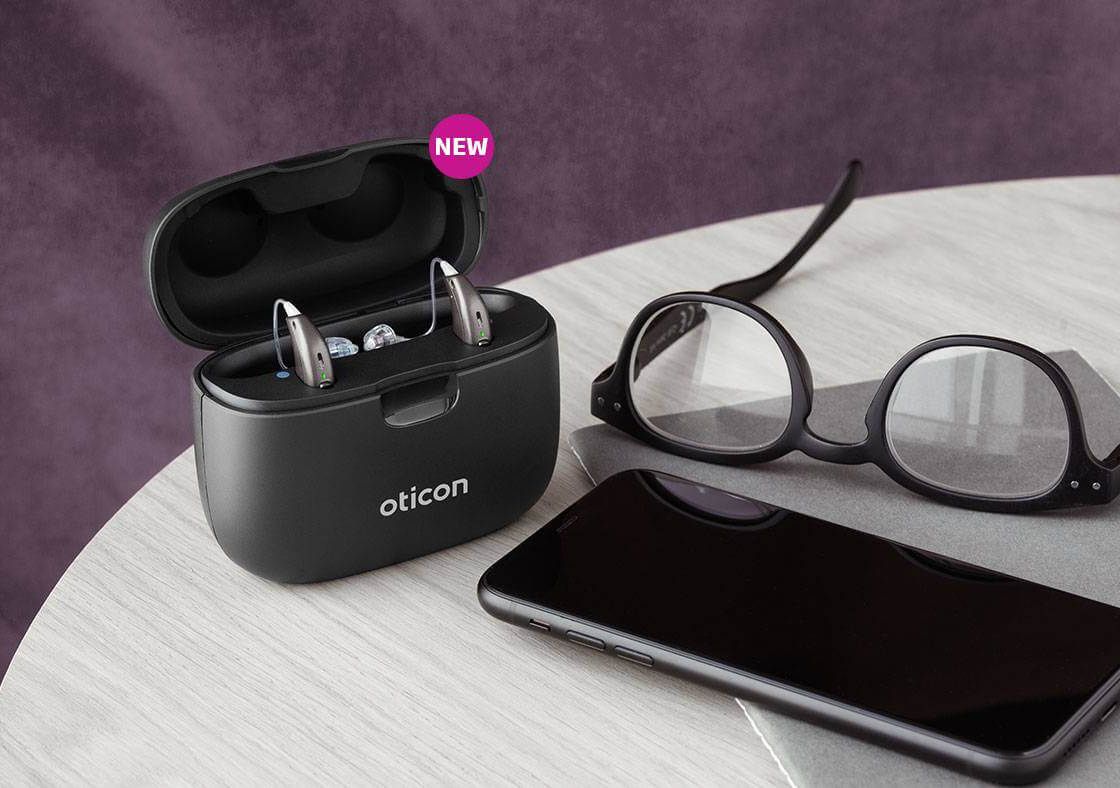A pair of Oticon Intent hearing aids beside a phone and reading glasses